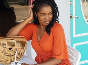 Photo of Smiling Woman in Orange V-neck Short-sleeved Dress Sitting Outside Store Holding Ice Cream While Looking Away